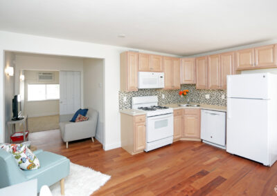 Glenbrook Centre Apartments. Cedar Rapids Iowa. Kitchen with view of living room and bedroom. Wood flooring. White walls. Wood cabinets. Gas range. Microwave. Dual stainless steel sink. Tile backsplash. Dishwasher. Refrigerator. Pocket door dividing living room space and bedroom. Carpeting in bedroom. Window with blinds in bedroom.